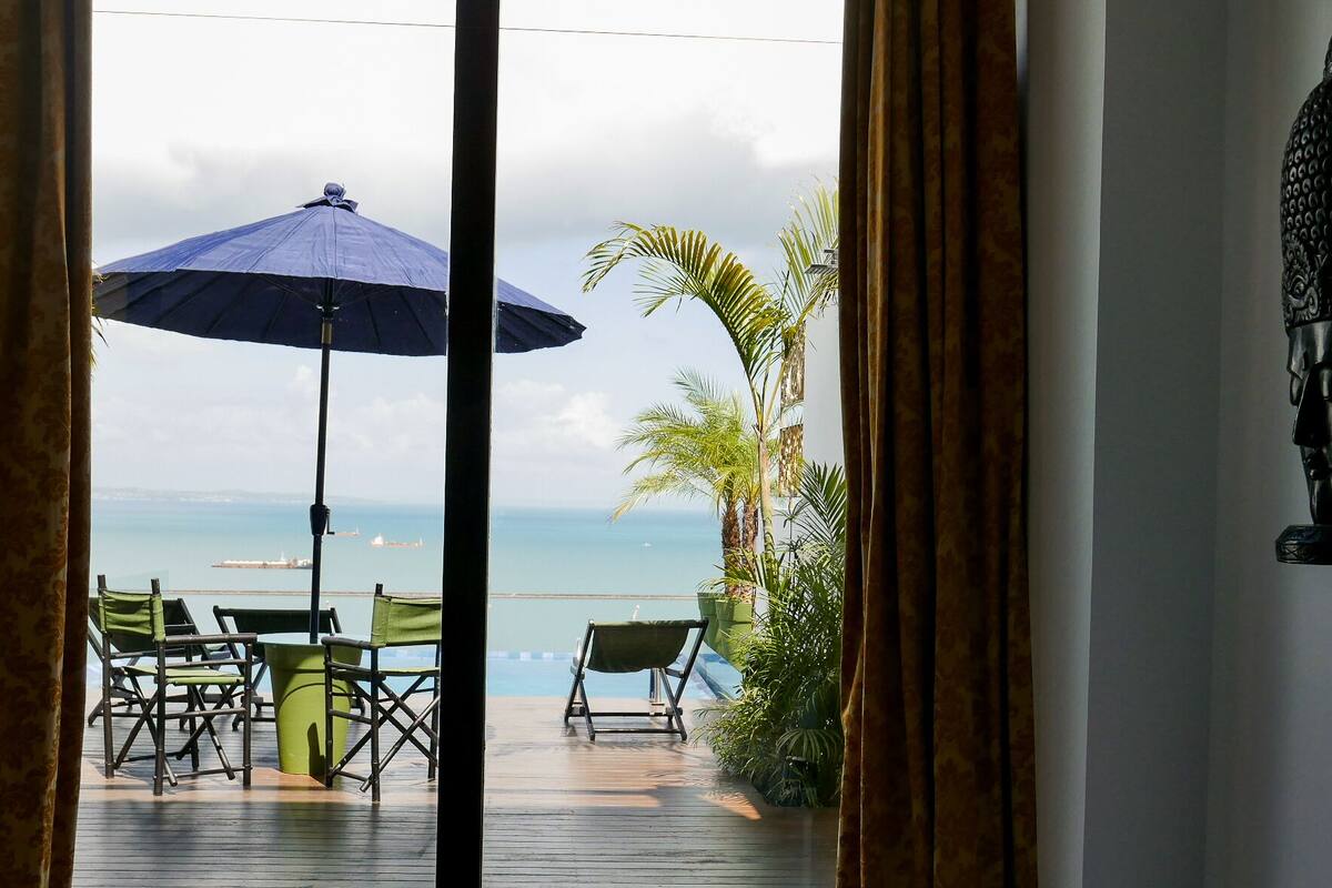 An outdoor seating area with a table and chairs, a blue umbrella, and a view of the ocean.