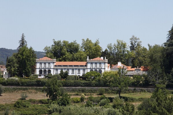 A large, historic building with multiple wings and a red-tiled roof, surrounded by lush greenery and trees.