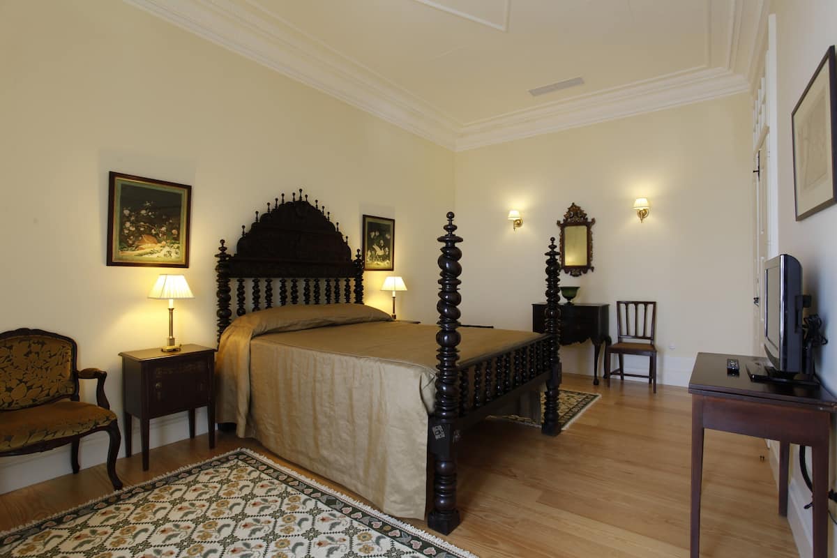 A traditional four-poster bed with a wooden frame, a matching nightstand, a chair, a television, and a wall-mounted mirror.