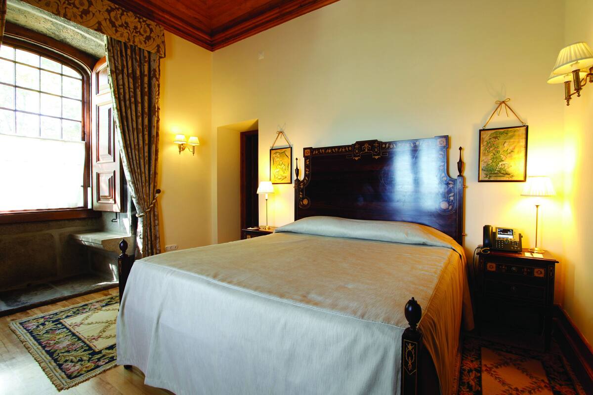 A well-lit hotel room with a large bed, a nightstand with a lamp, a wooden headboard, and a window with curtains.