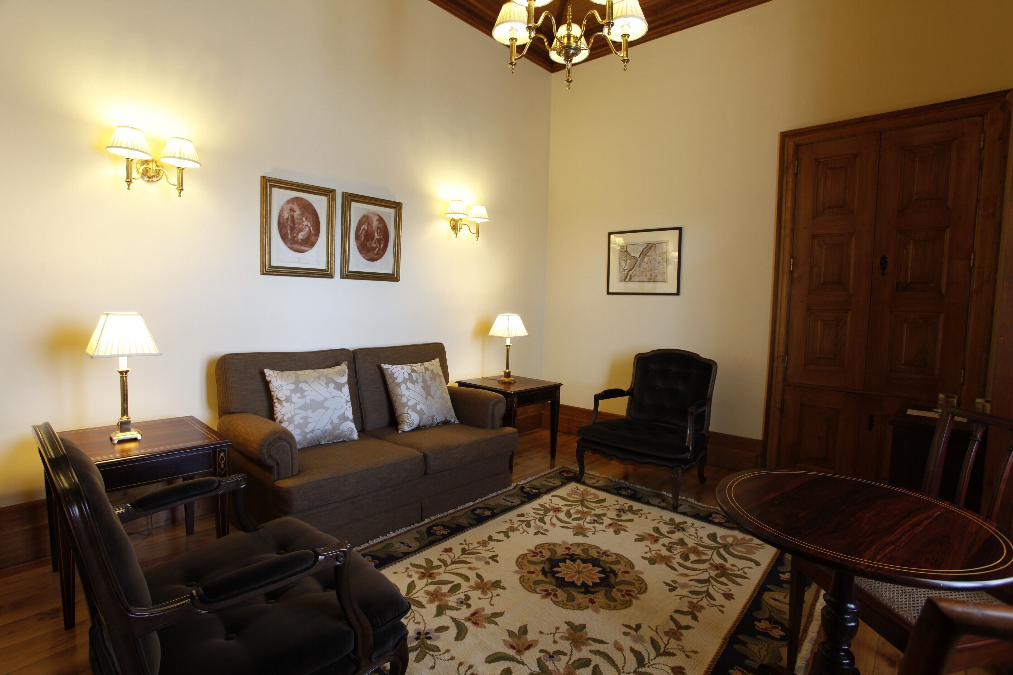 Una sala de estar con sofá, dos sillones, una mesa de centro y una mesa de comedor. Hay lámparas de pared, cuadros enmarcados y una puerta de madera.