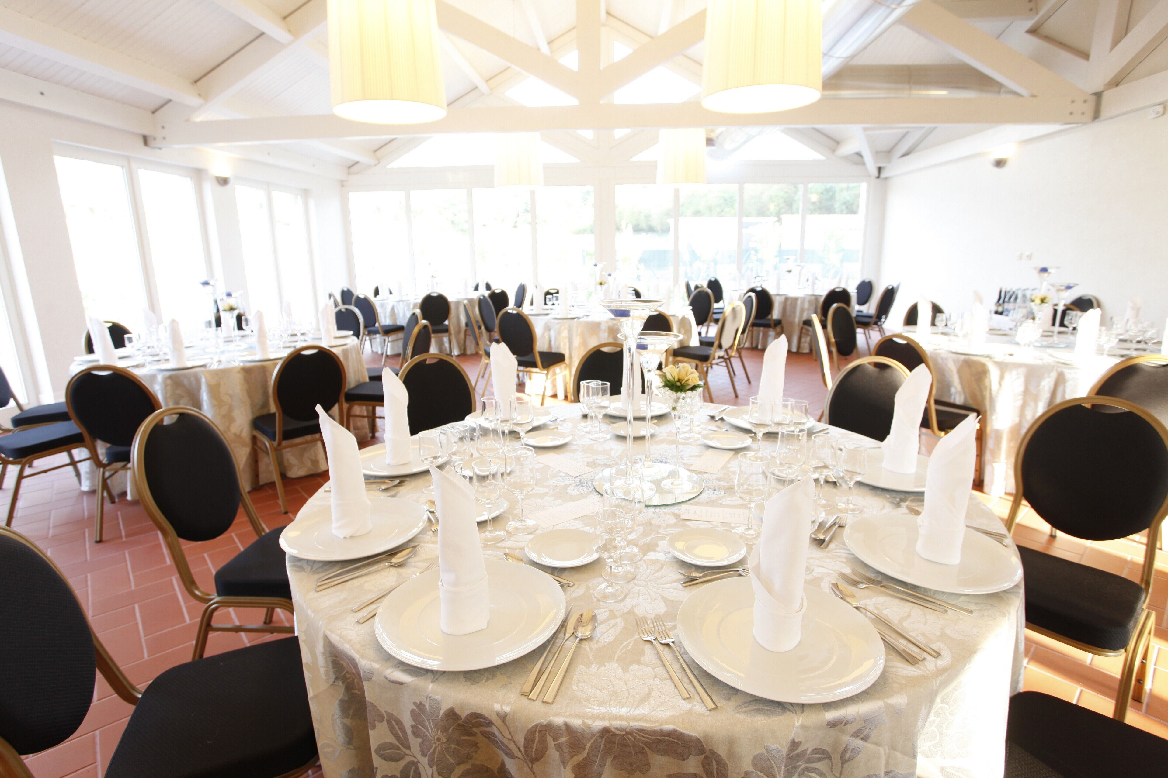 A banquet hall with round tables set for a formal event, featuring white tablecloths, gold cutlery, and black chairs.