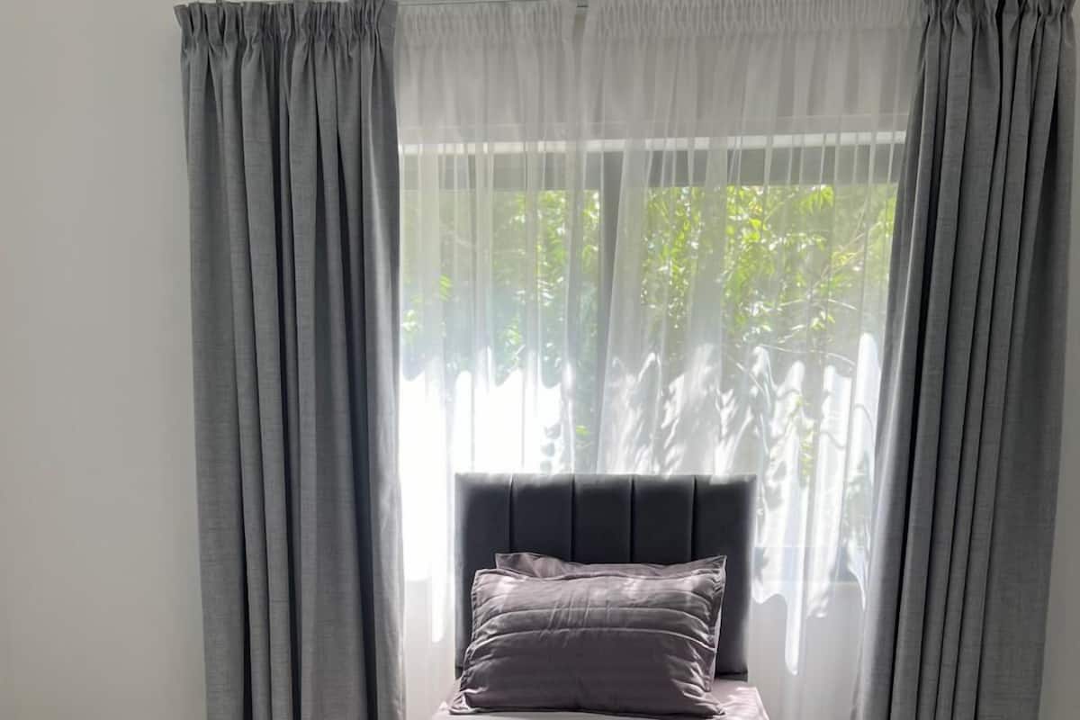 A single bed with a grey headboard and matching pillows, set against a window with sheer grey curtains.