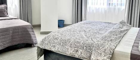 A bedroom with two beds, a large window with curtains, a wall-mounted air conditioner, and a blue laundry basket.