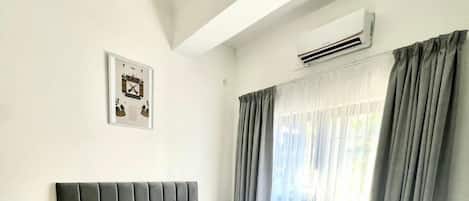 Un dormitorio con una cama grande, cabecero, mesita de noche, aire acondicionado instalado en la pared y una ventana con cortinas.