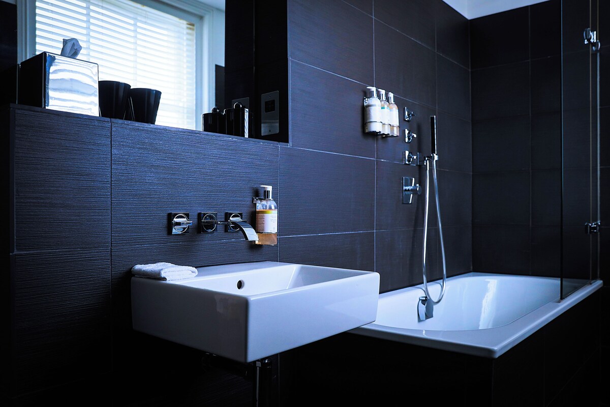 Un baño moderno con un lavamanos blanco, ducha y paredes de azulejos oscuros.