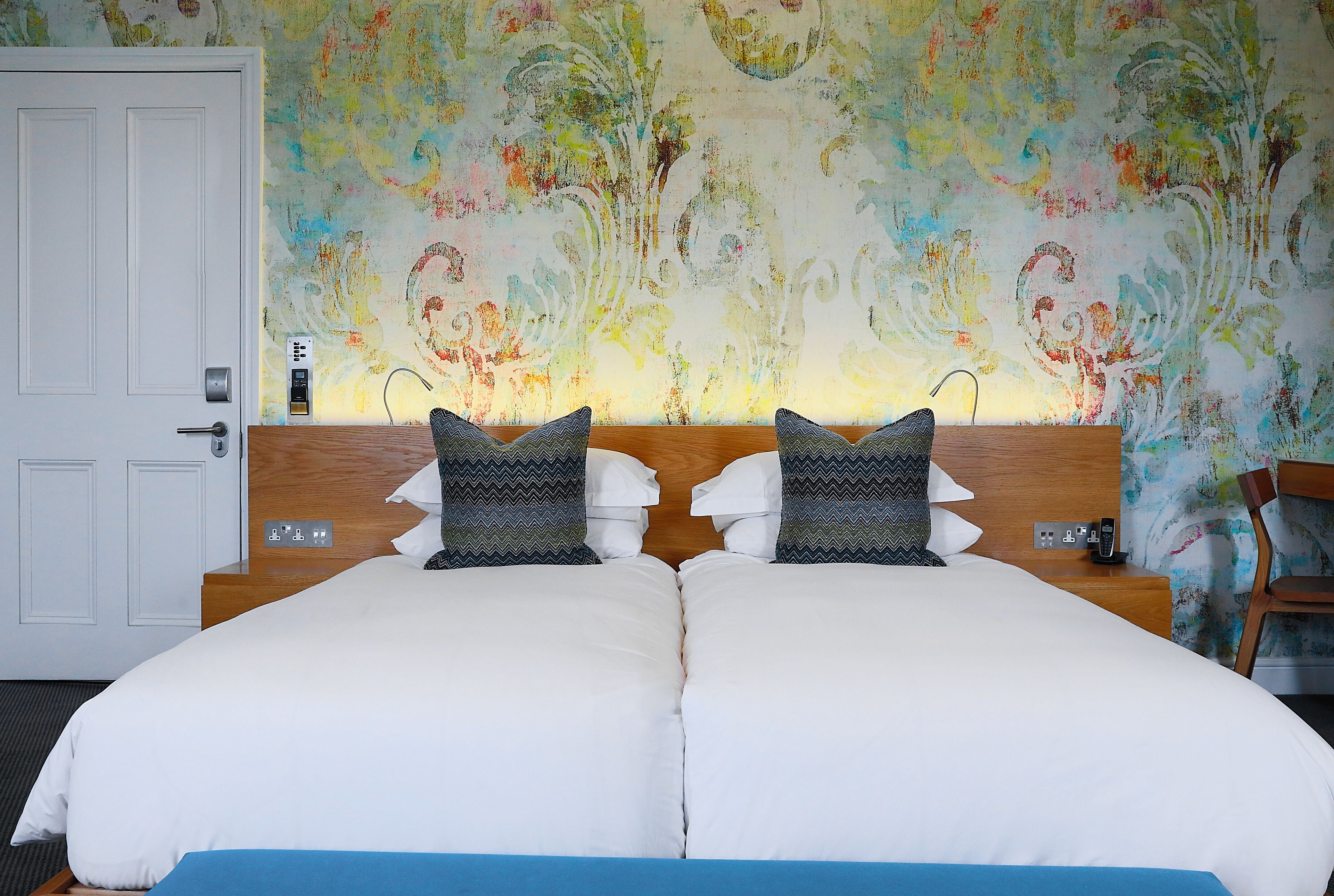 A double bed with white and blue bedding, a wooden headboard, and a colorful abstract wall mural.