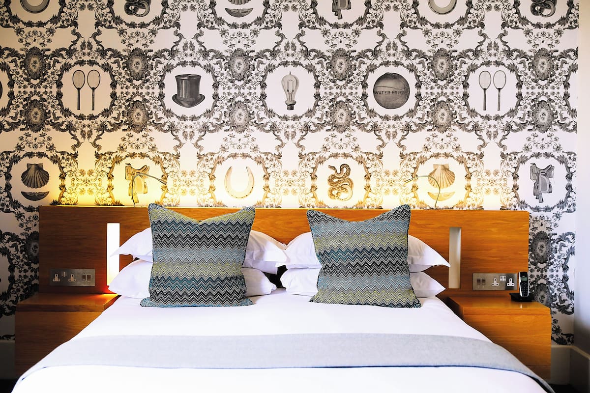A neatly made bed with patterned pillows, a wooden headboard, and a wall with decorative wallpaper.