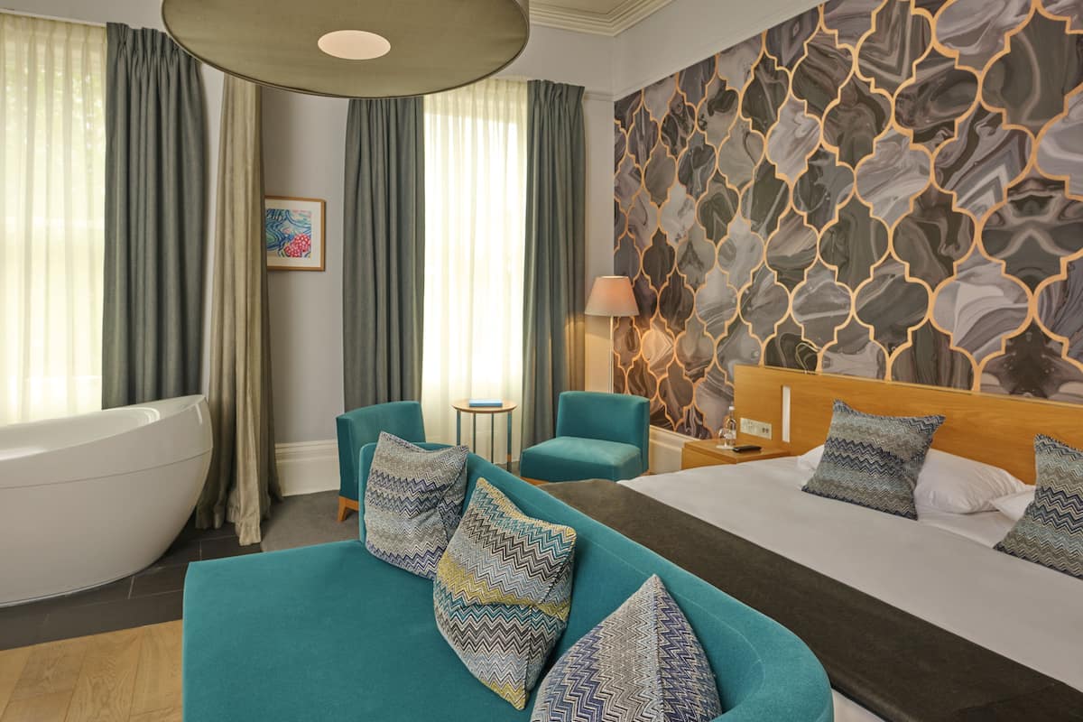 A modern hotel room with a large bed, a sofa, a bathtub, and a patterned wall.