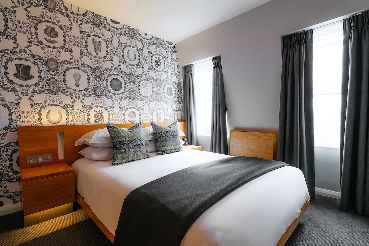 A neatly made bed with a black throw, a wooden nightstand, and a wall with a patterned wallpaper.