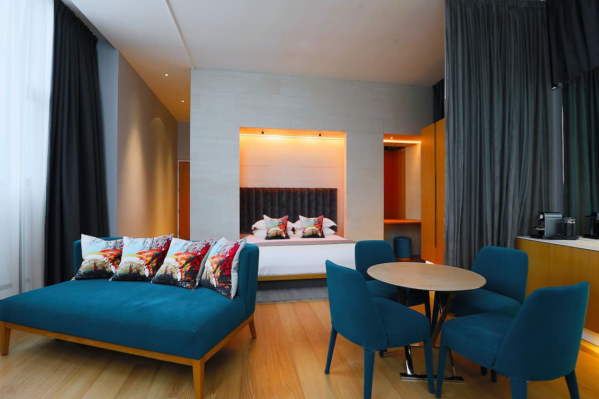 A modern hotel room with a bed, a sofa, a dining table, and chairs.