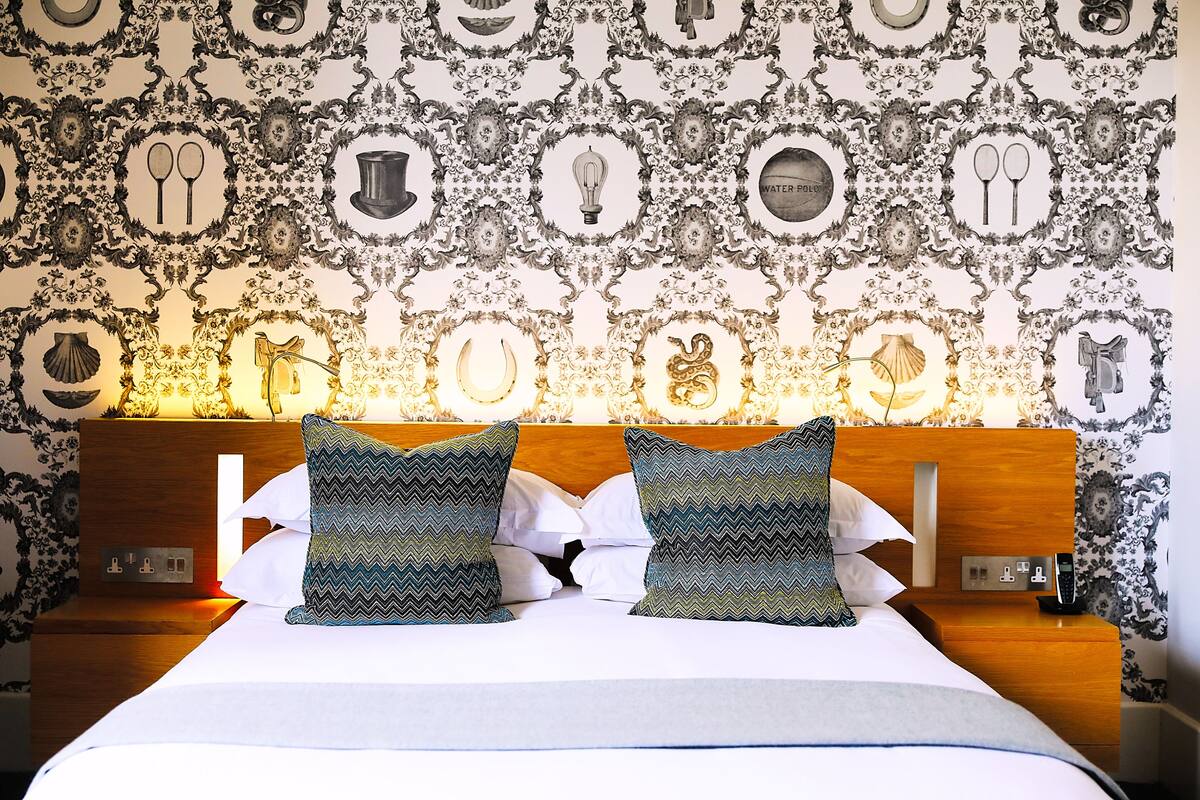 A neatly made bed with patterned pillows, a wooden headboard, and a wall with decorative wallpaper.