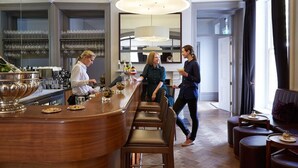 A bar area with a wooden counter, bar stools, and a large mirror. There are wine glasses and bottles on the shelves behind the counter. - Malmaison Cheltenham (Cheltenham)