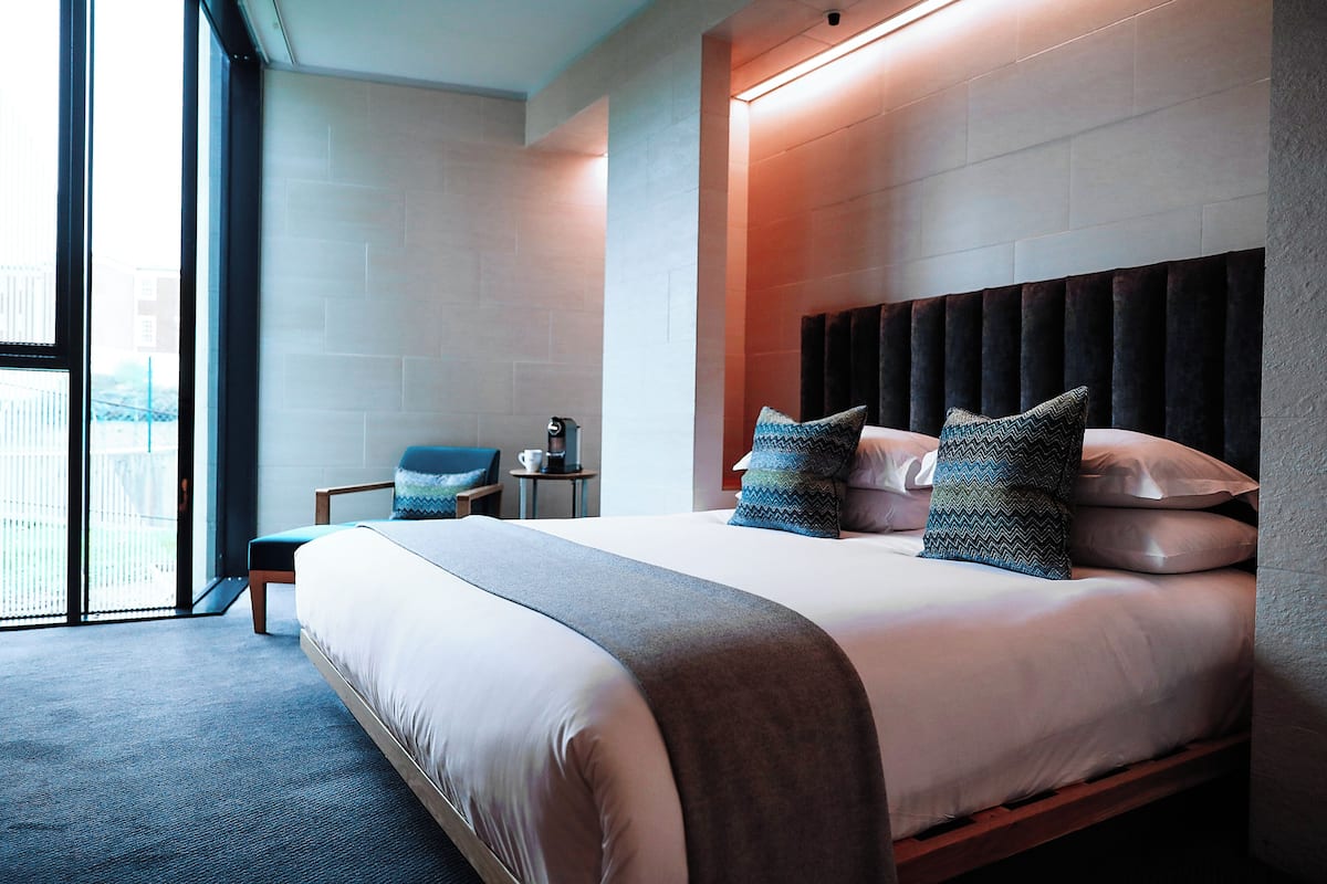 A modern hotel room with a large bed, a seating area with a chair, and a view through a large window.