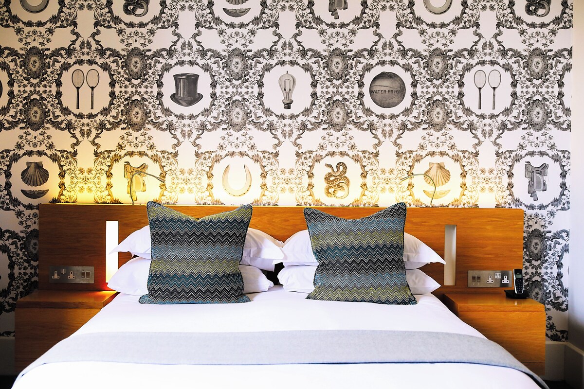 A neatly made bed with patterned pillows, a wooden headboard, and a wall with decorative wallpaper.