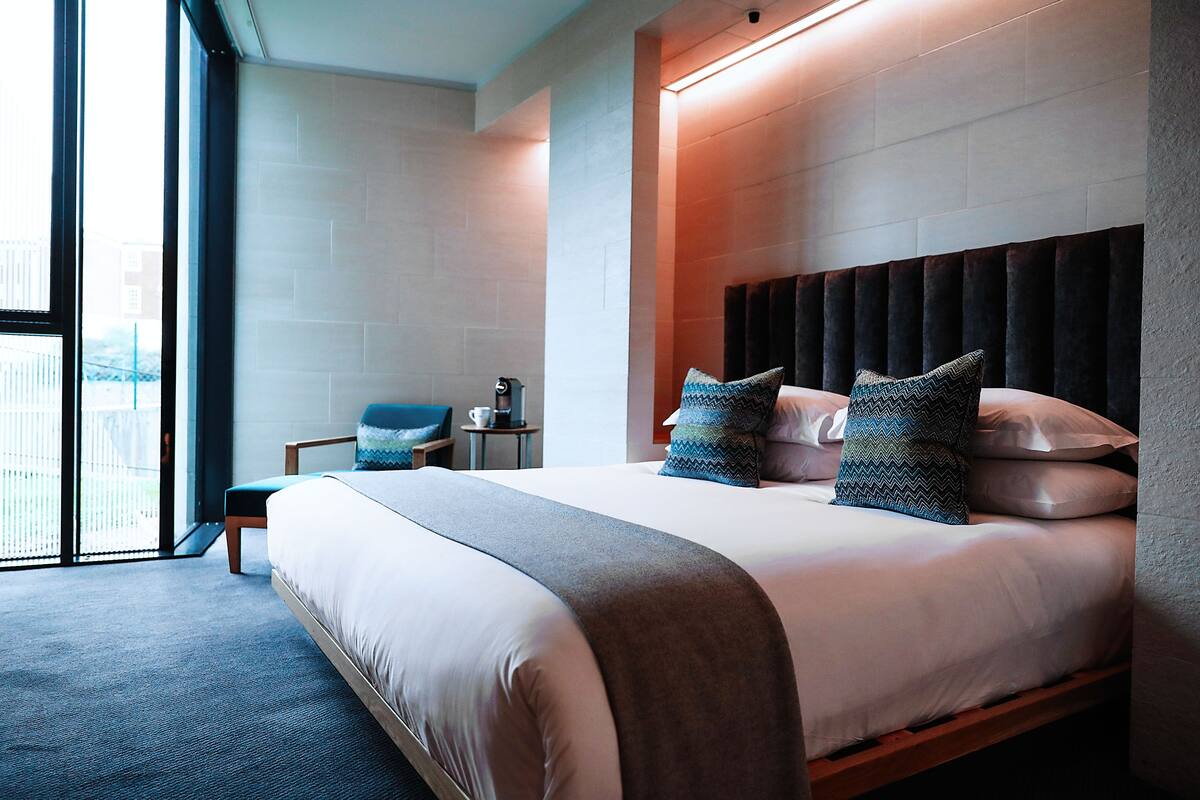 A modern hotel room with a large bed, a seating area with a chair, and a view through a large window.