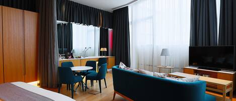 Ein modernes Hotelzimmer mit Bett, einer Couch, einem Fernseher, einer Essecke und einer Küche im Hintergrund.