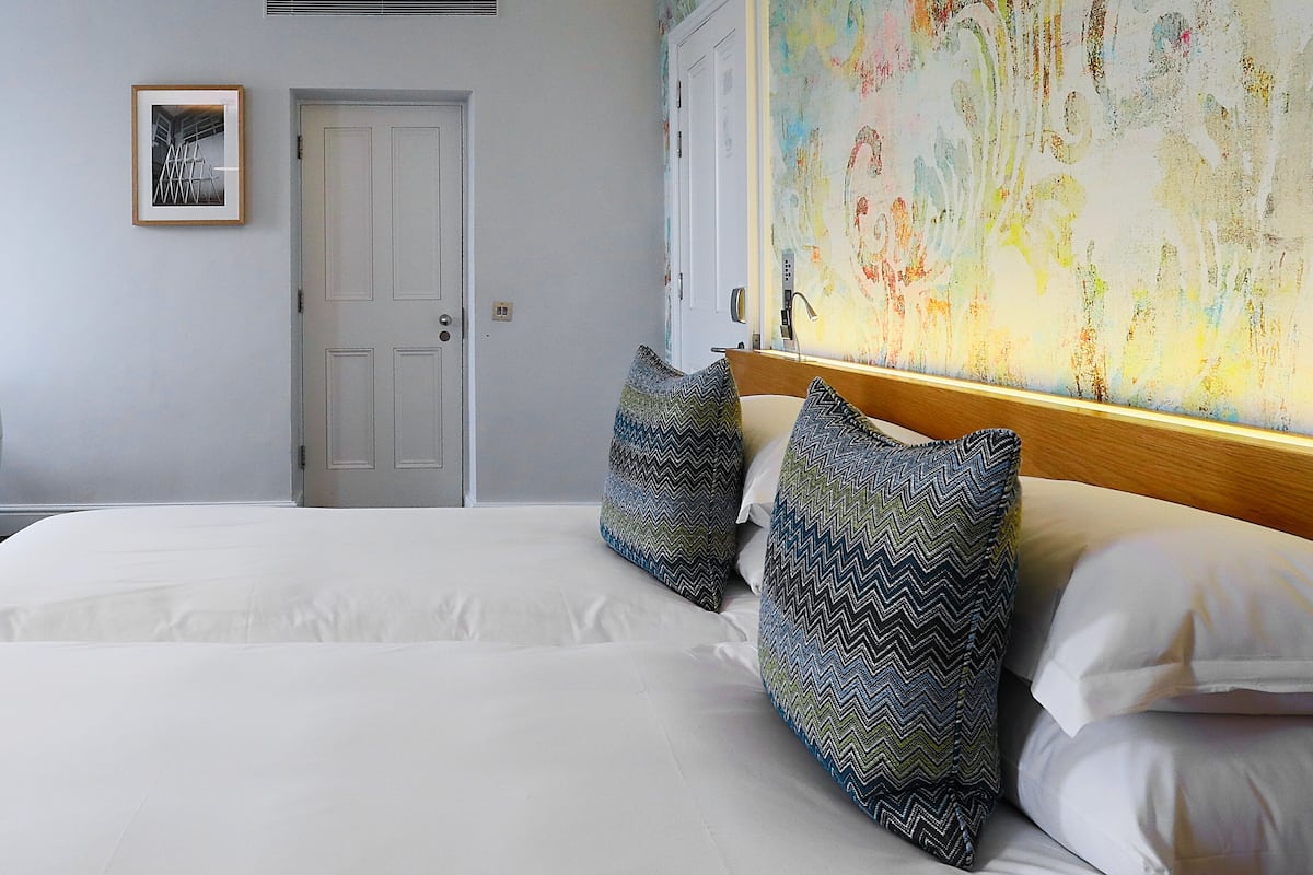 A hotel room with a large bed, two pillows, a door, a framed picture, and a colorful abstract painting on the wall.
