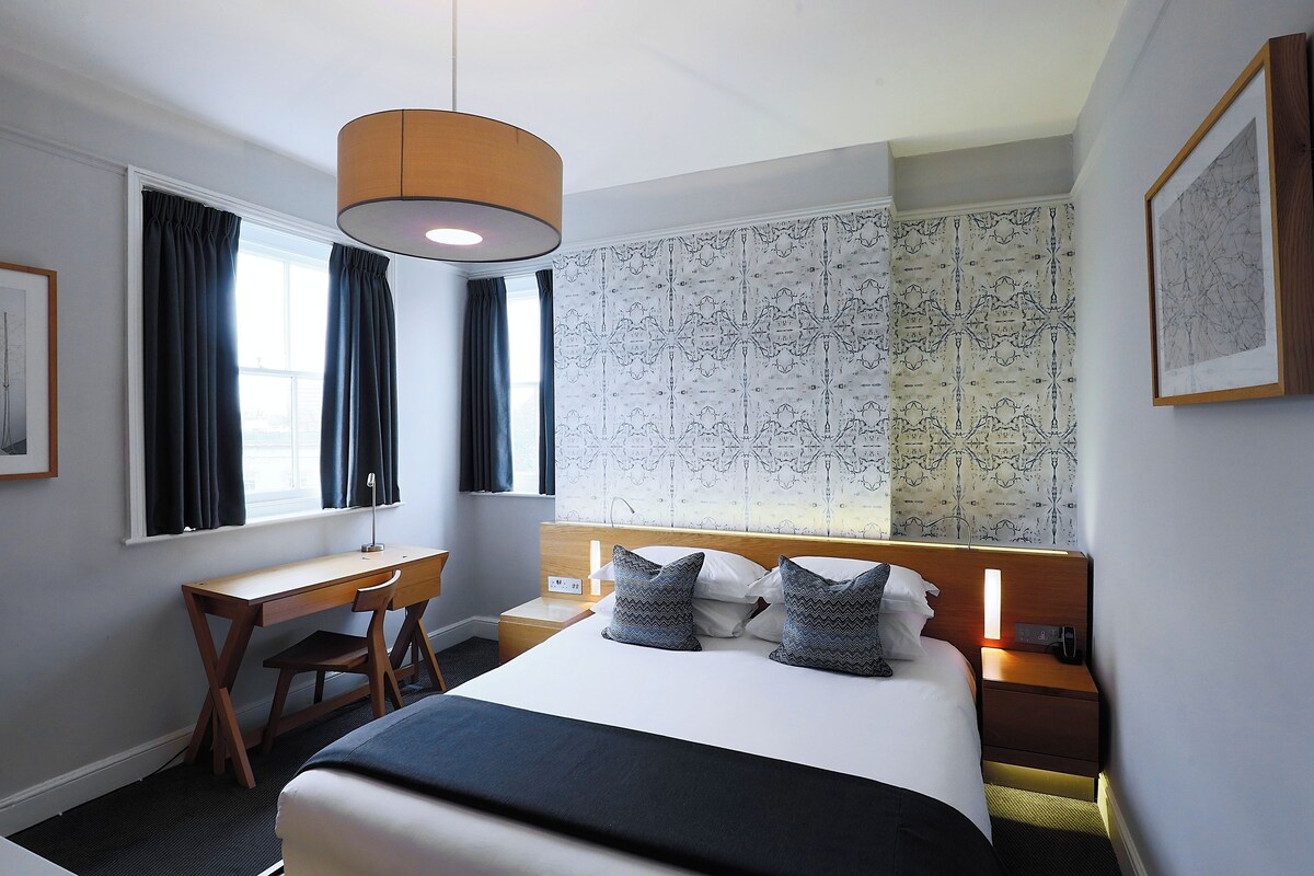 A hotel room with a bed, desk, chair, and a large window with curtains.