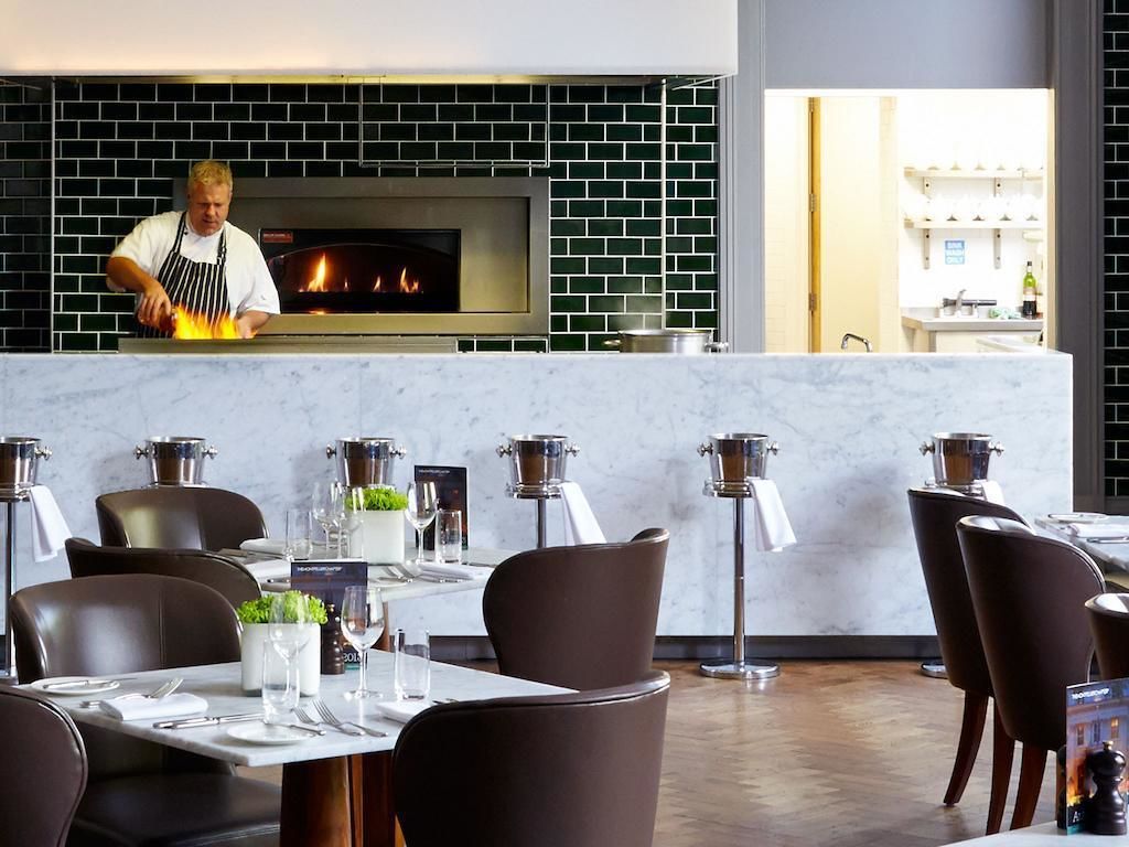 En restaurang med en tegelvägg och en vedeldad ugn. Borden är dukade med vita dukar och det står bruna stolar runt dem.