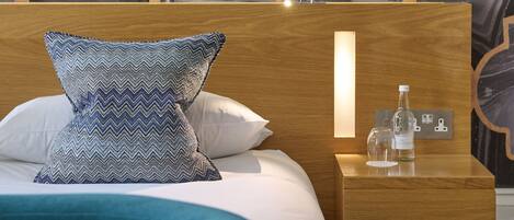 A neatly made bed with patterned pillows, a wooden headboard, and a bedside table with a bottle and glass.
