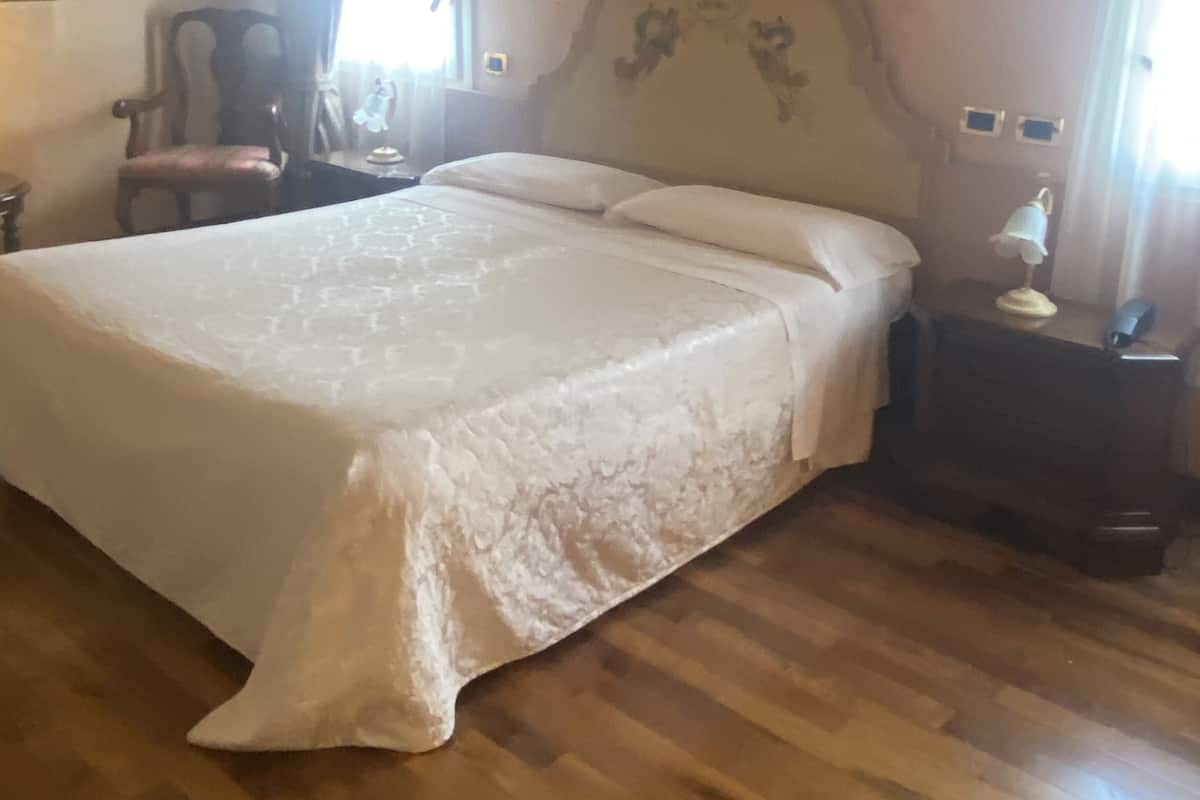 A bedroom with a large bed, wooden flooring, a chair, a nightstand, and a painting on the wall.