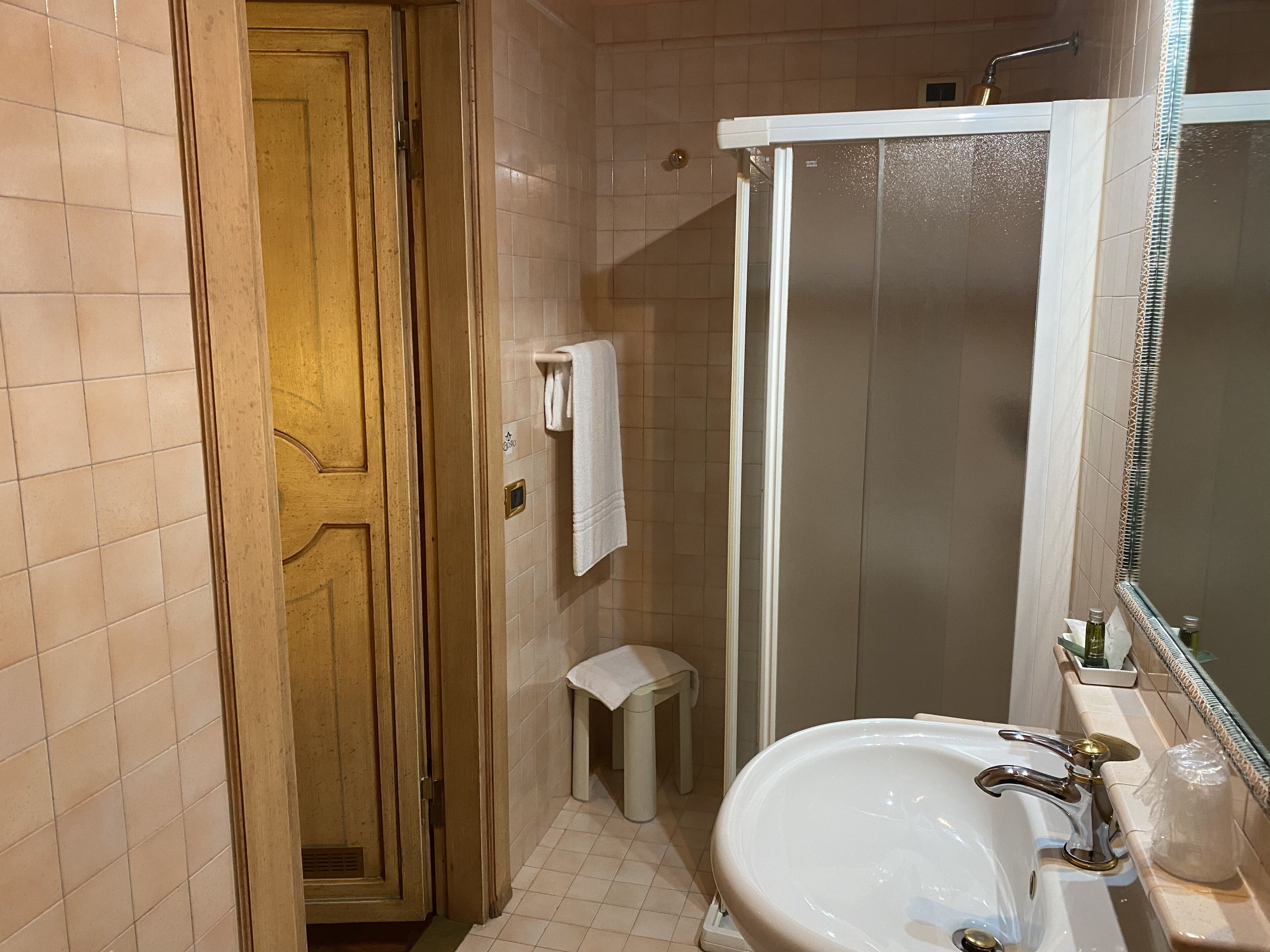 A bathroom with a wooden door, a white sink, a glass shower enclosure, and a small stool.