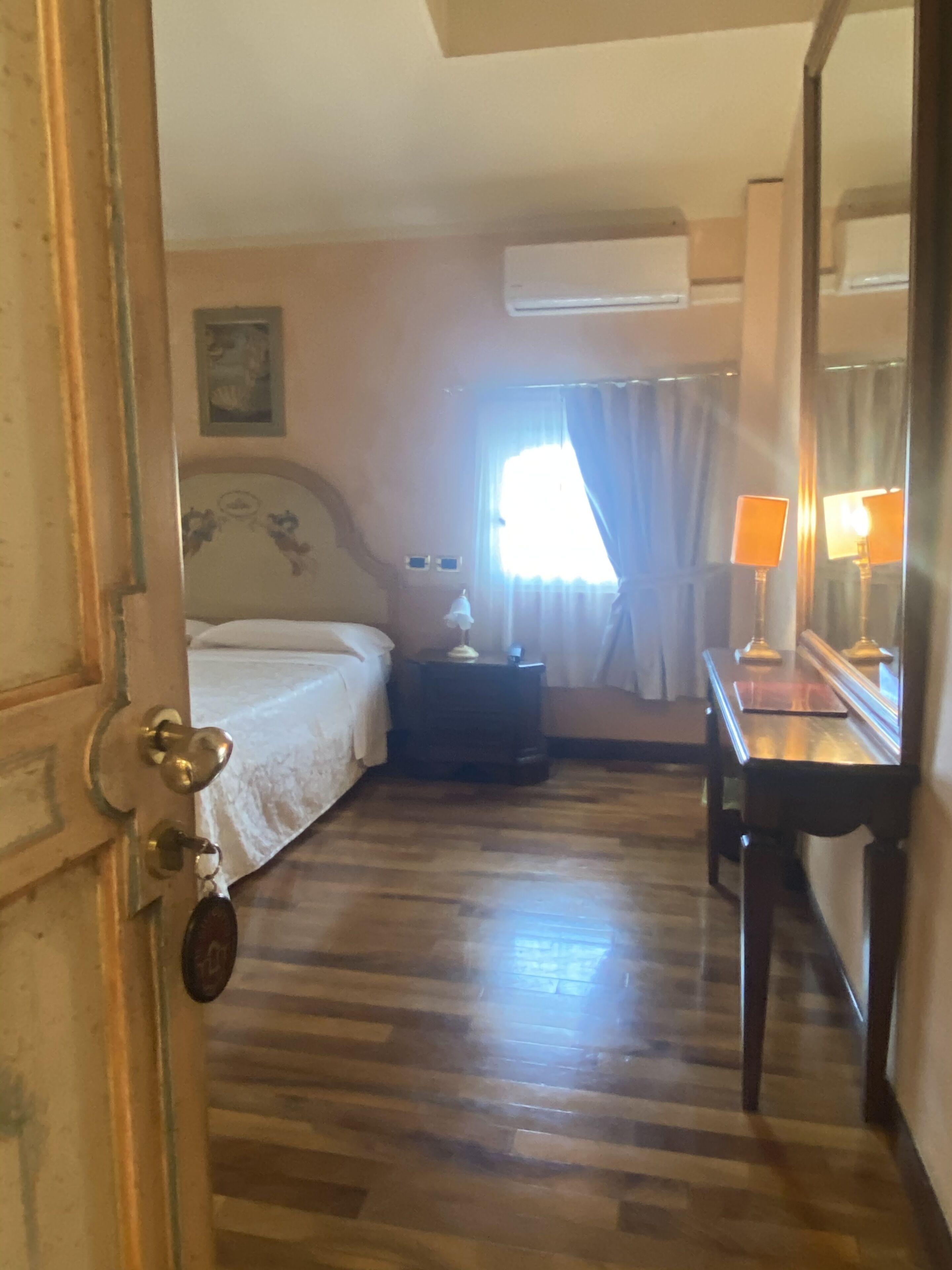 Una habitación de hotel con piso de madera, una cama con cabecera floral, una mesita de noche con lámpara y un espejo grande.