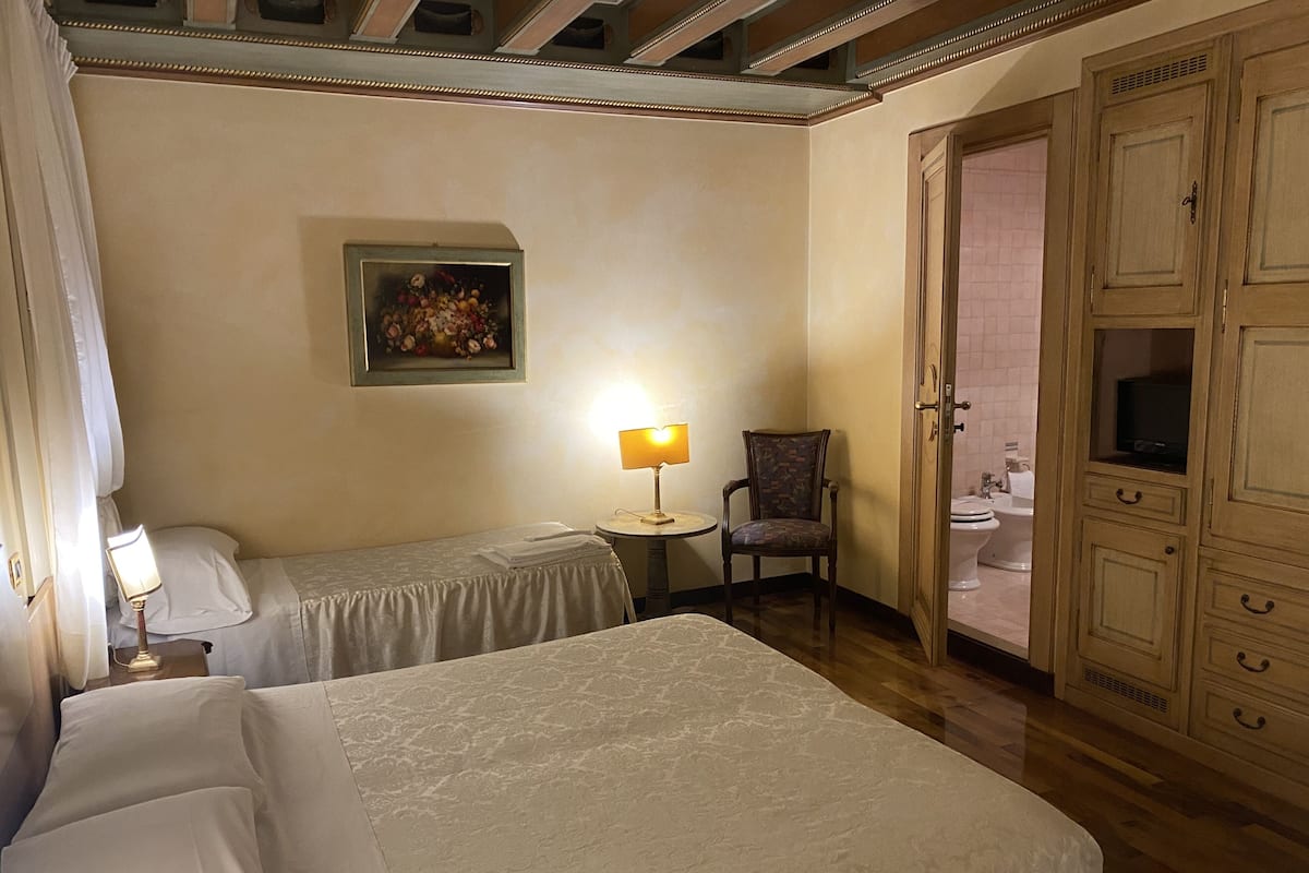 A hotel room with two beds, a chair, a table with a lamp, a painting on the wall, and a bathroom visible through an open door.