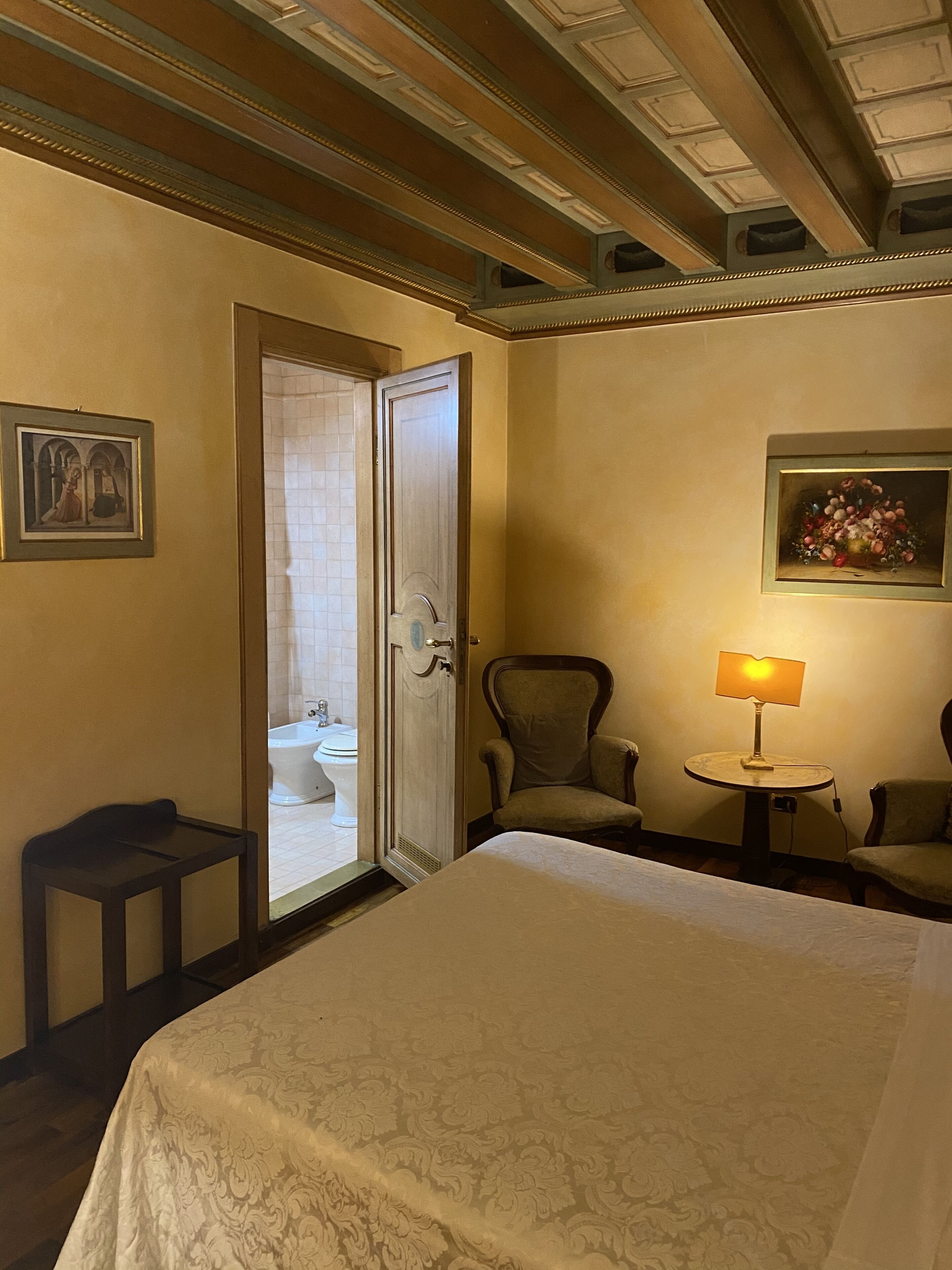 Quarto de hotel com uma cama, uma cadeira, uma pequena mesa com abajur e uma porta para o banheiro.