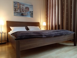 Una cama bien hecha con un cabecero oscuro y almohadas blancas, un cuadro de un paisaje urbano encima, y una cortina con estampado floral a la derecha.