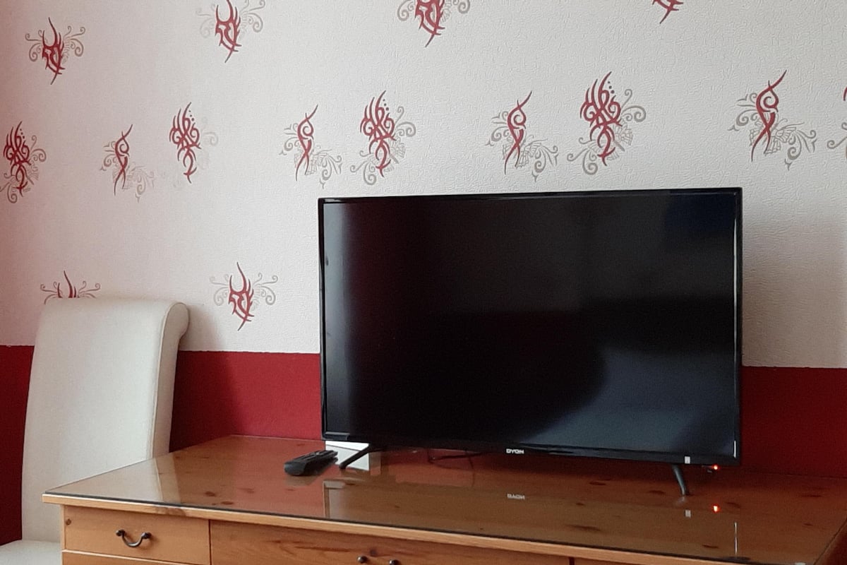Un televisor sobre un escritorio de madera con cajones, frente a una pared con papel tapiz de estampado rojo y blanco.