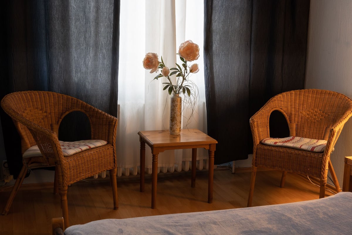 Dos sillas de mimbre con asientos acolchados, una pequeña mesa de madera con un jarrón de flores y una ventana con cortinas oscuras.
