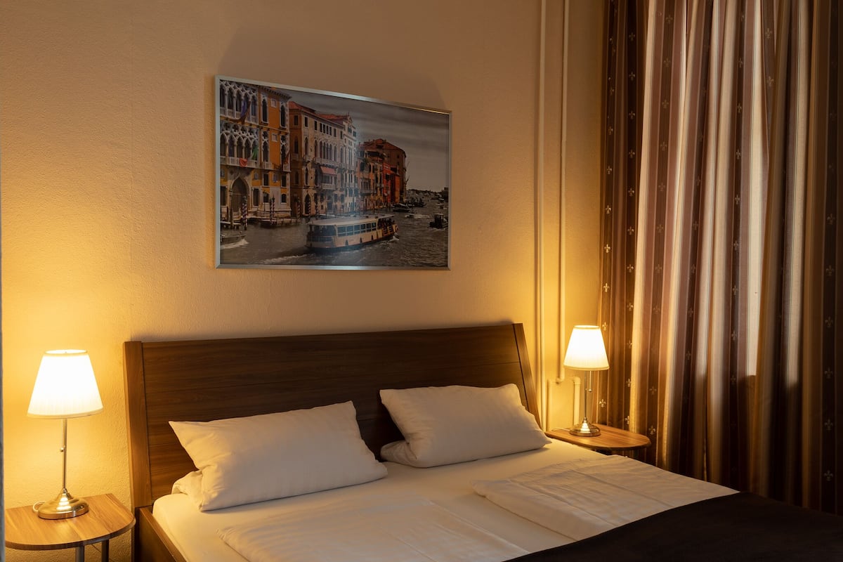 Una habitación de hotel con una cama, dos lámparas de noche, un cuadro de un paisaje urbano y una cortina.