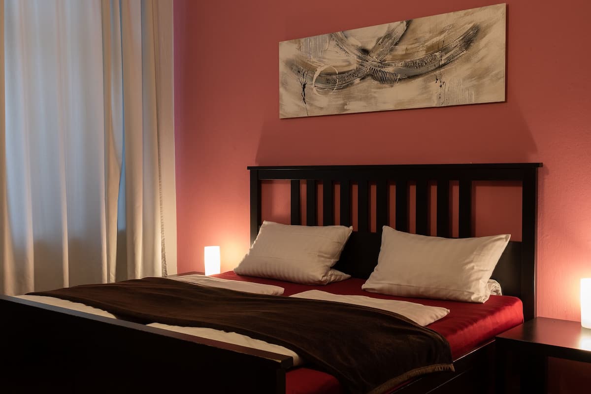 Una cama bien hecha con una colcha roja y dos almohadas blancas, un cabecero de madera oscura y un cuadro en la pared.