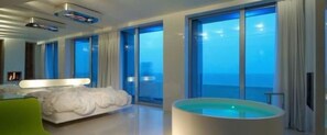 Una habitación de hotel moderna con una cama grande, una bañera y vista al océano a través de ventanales de piso a techo.