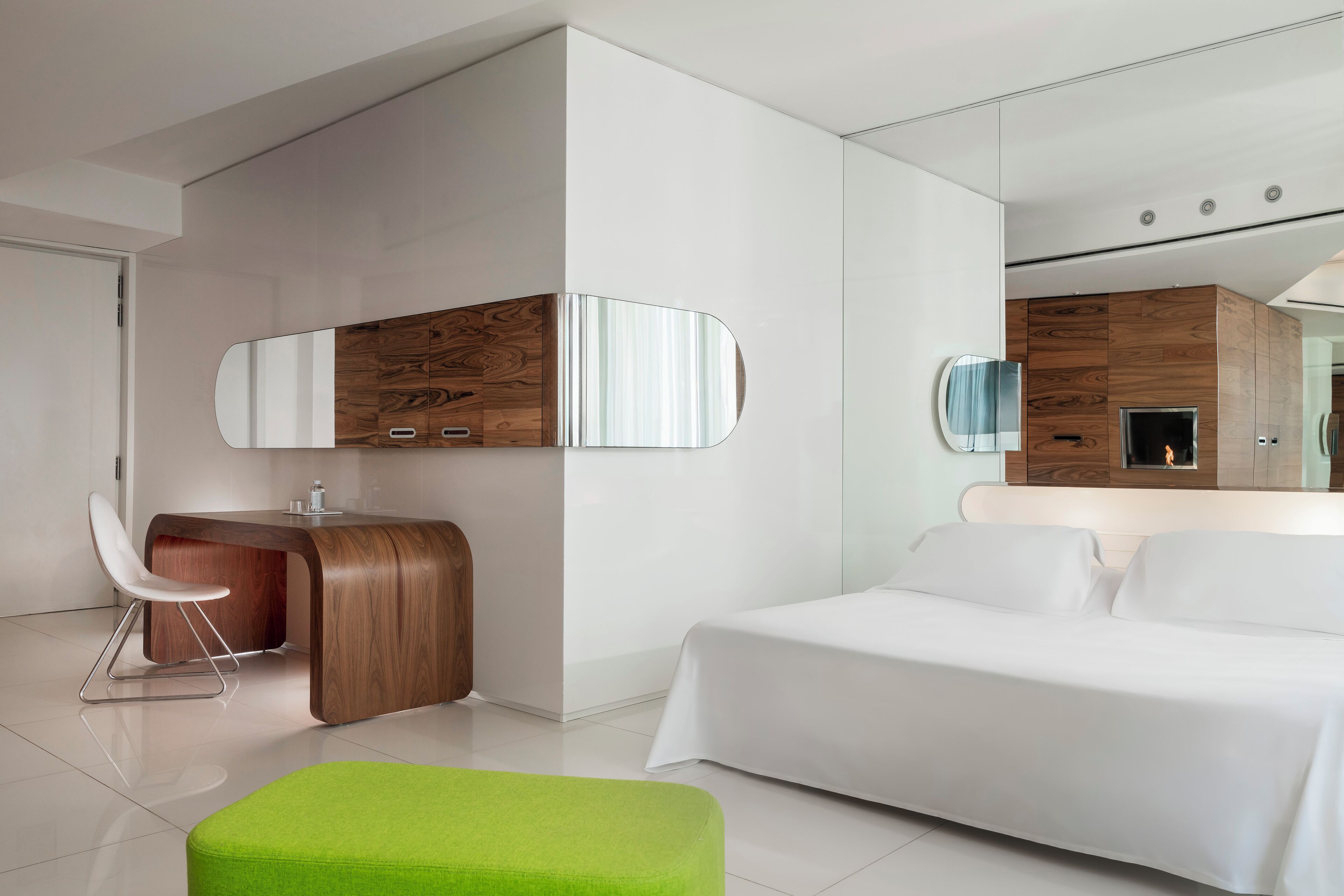 A modern hotel room with a bed, a green ottoman, a wooden vanity with a mirror, and a fireplace.