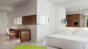 A modern hotel room with a bed, a green ottoman, a wooden vanity with a mirror, and a fireplace.