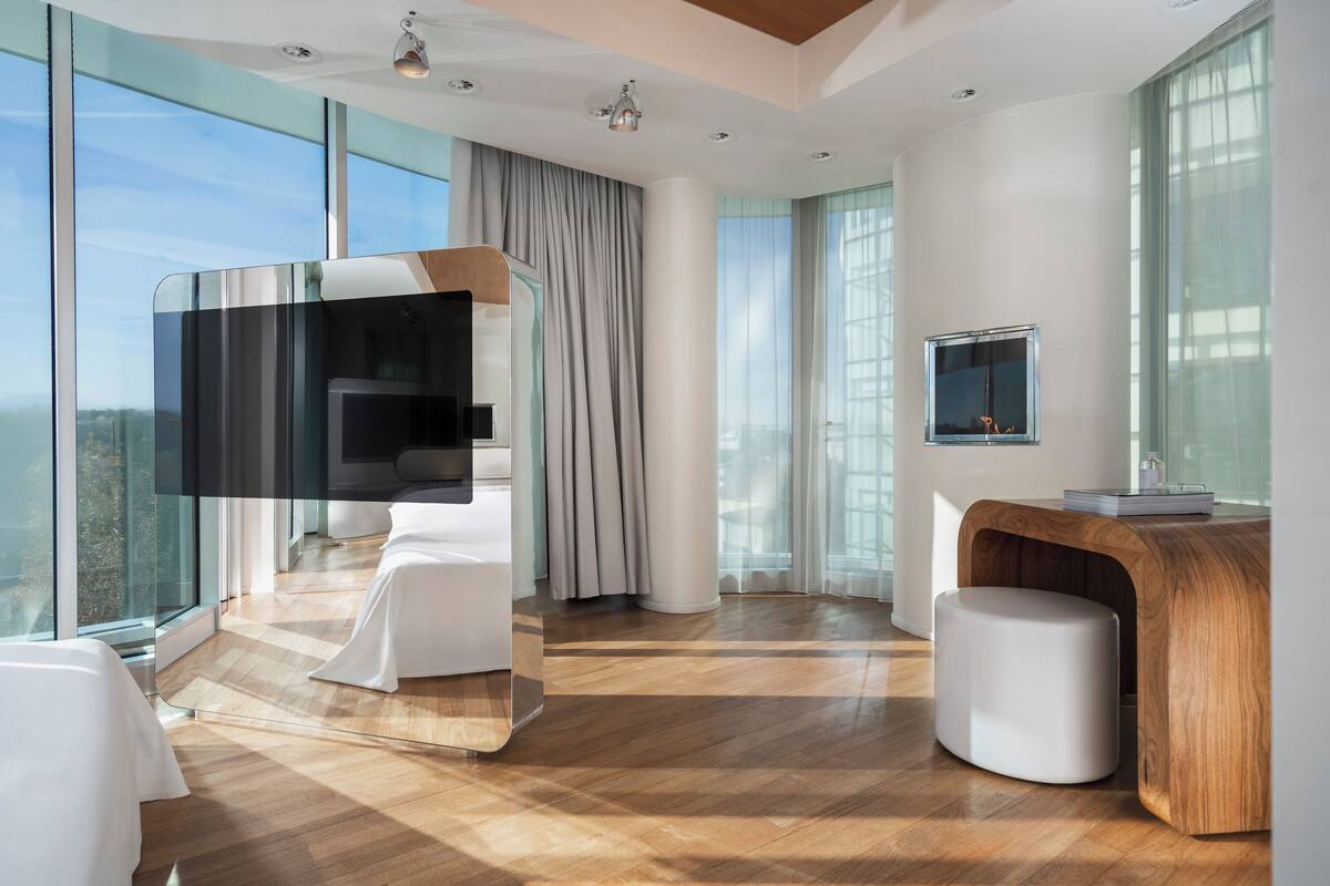 A modern hotel room with a large window, a wooden desk, a TV, and a white bed.