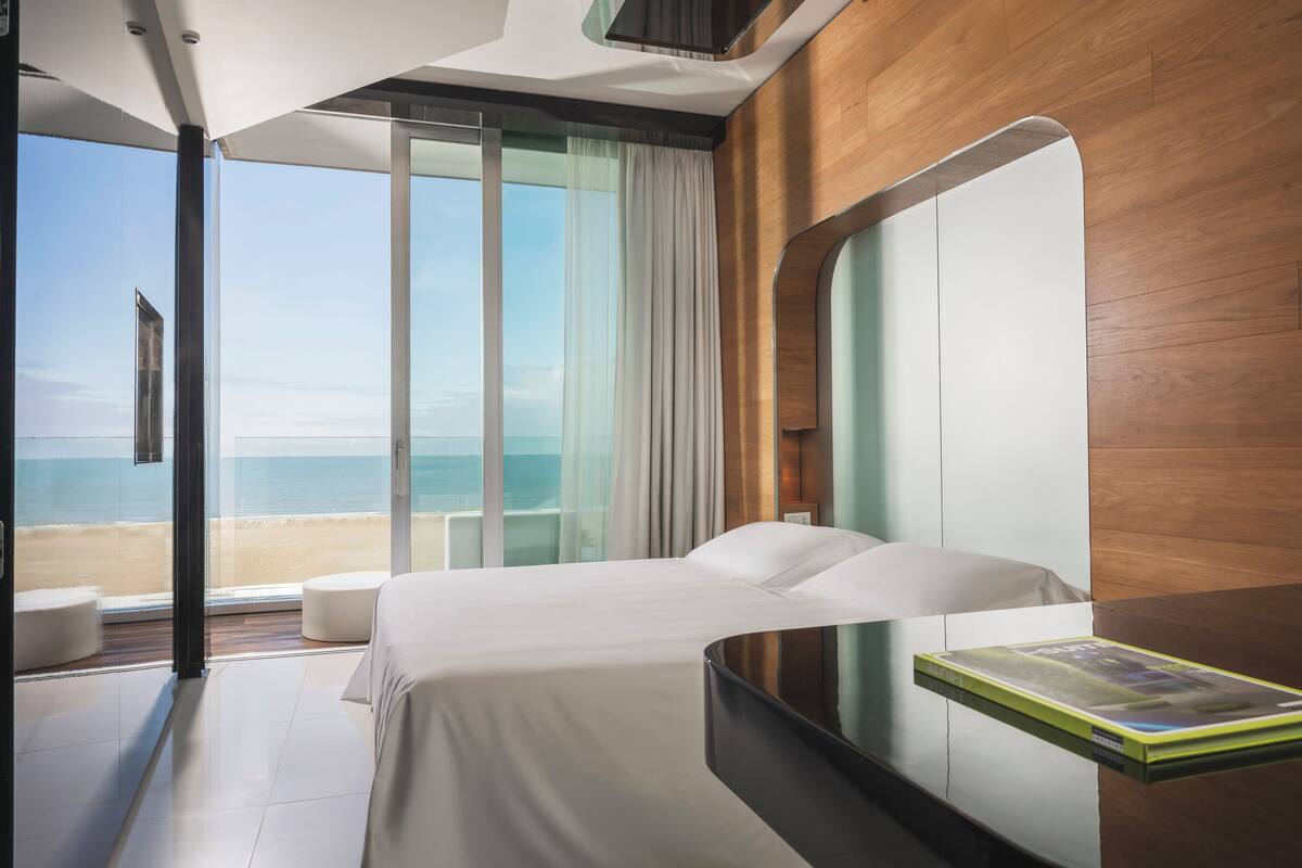 A modern hotel room with a large bed, a bedside table with a book, and a view of the beach.