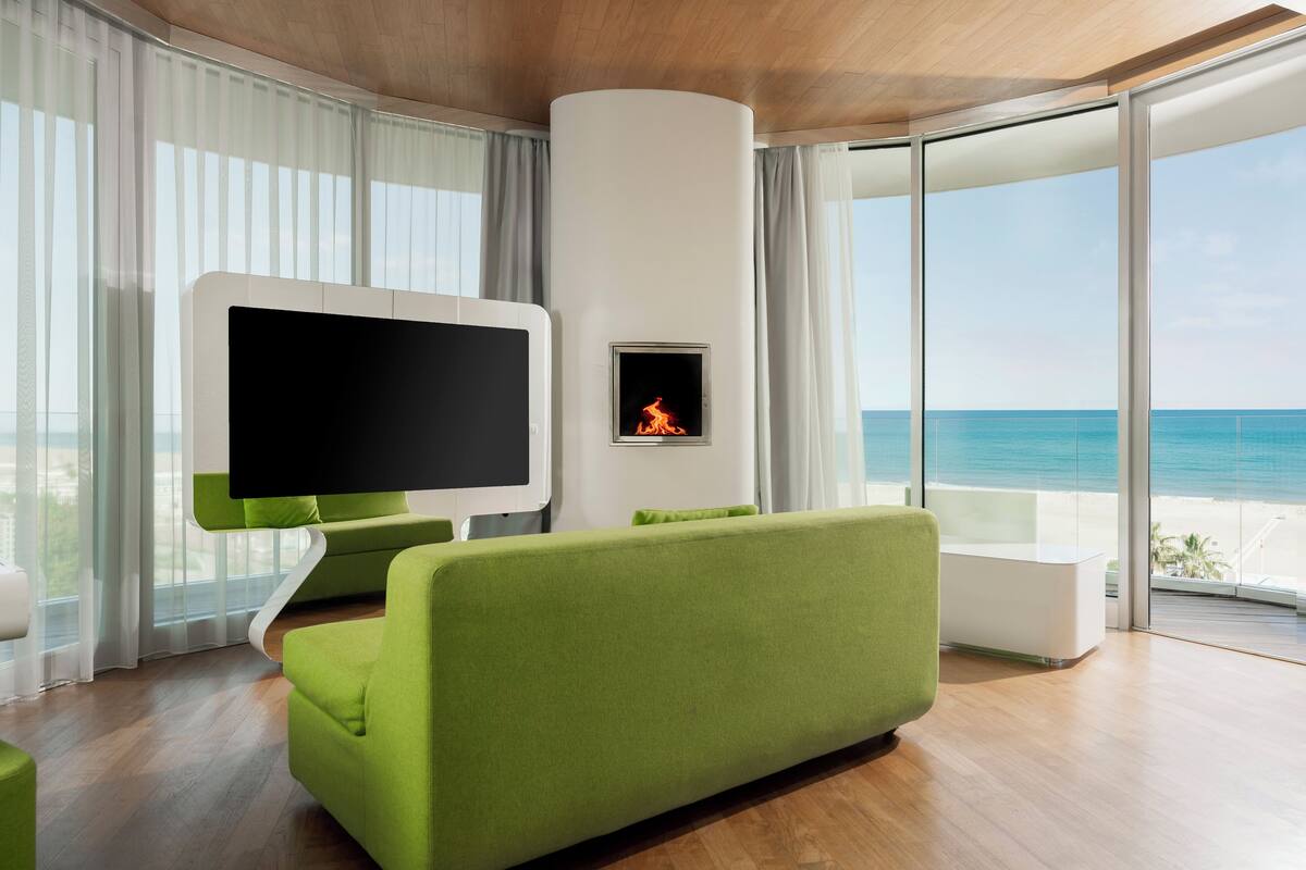 A modern living room with a large flat-screen TV, a fireplace, a green sofa, and a balcony with a view of the ocean.