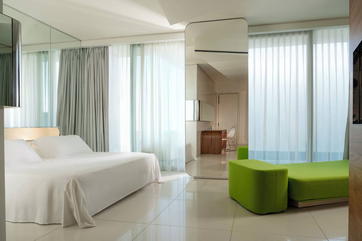 A modern hotel room with a large bed, a green ottoman, and a view of the city through large windows.