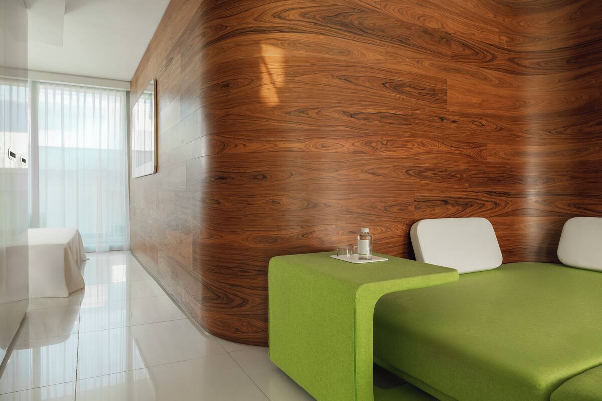 A modern hotel room with a curved wooden wall, a green bed, and a small table with a bottle and glass.