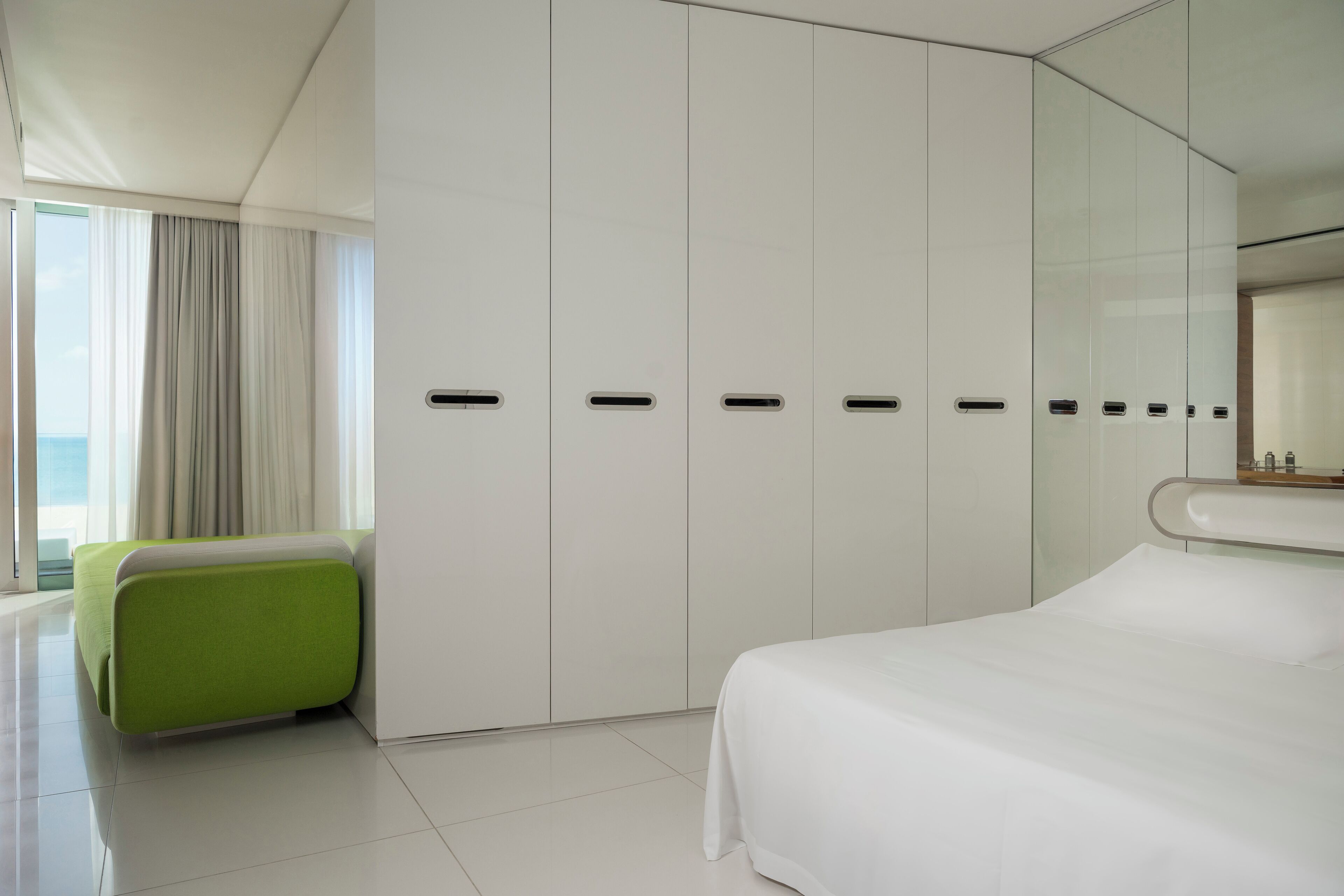 A modern hotel room with a large bed, a green accent chair, a wardrobe with multiple compartments, and a large window offering a view.