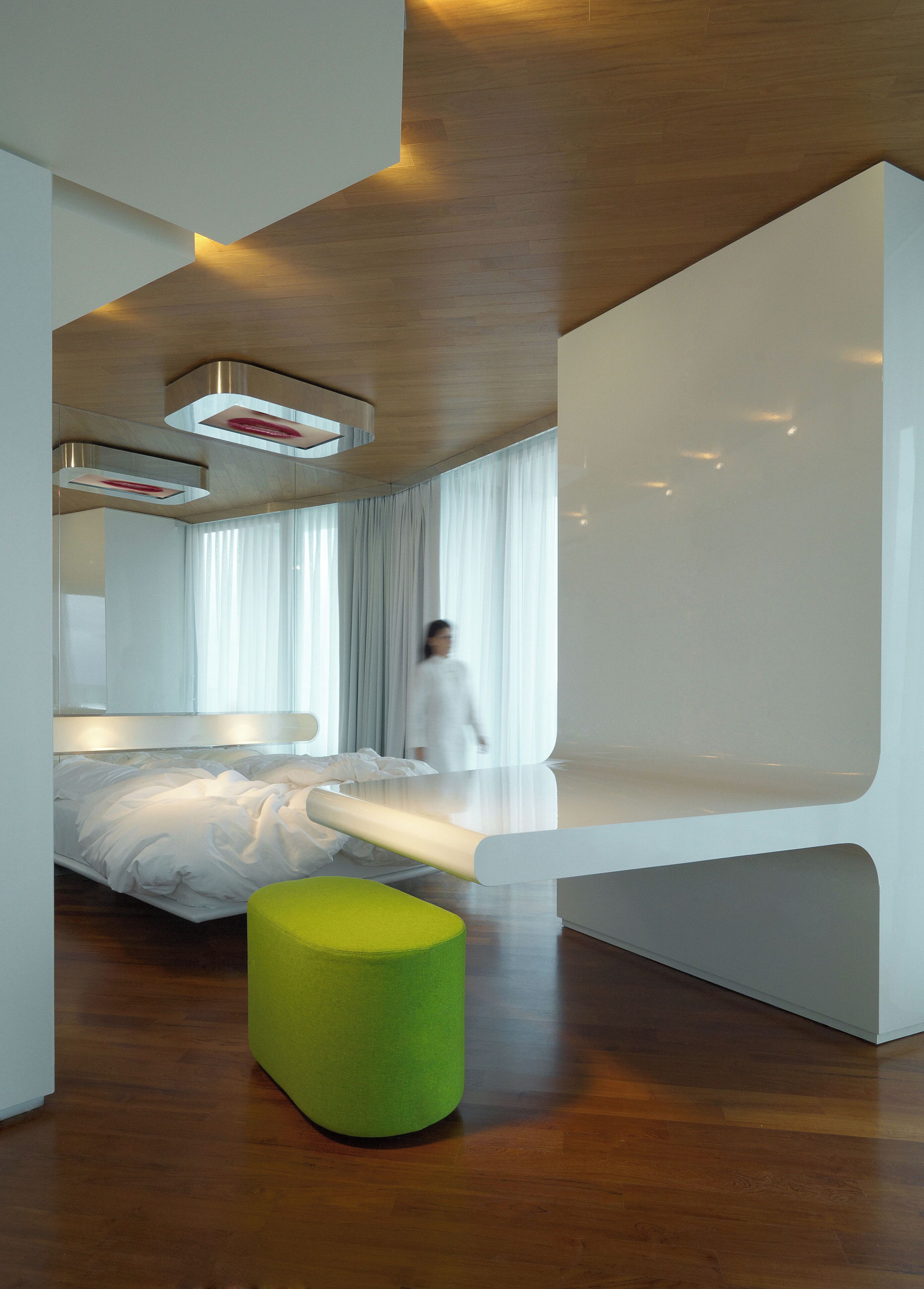 Ein modernes Schlafzimmer mit einem Bett, einem grünen Hocker und einer Person im Hintergrund.