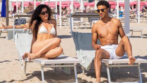 Two people sitting on lounge chairs at a beachside resort with umbrellas and beach chairs in the background.