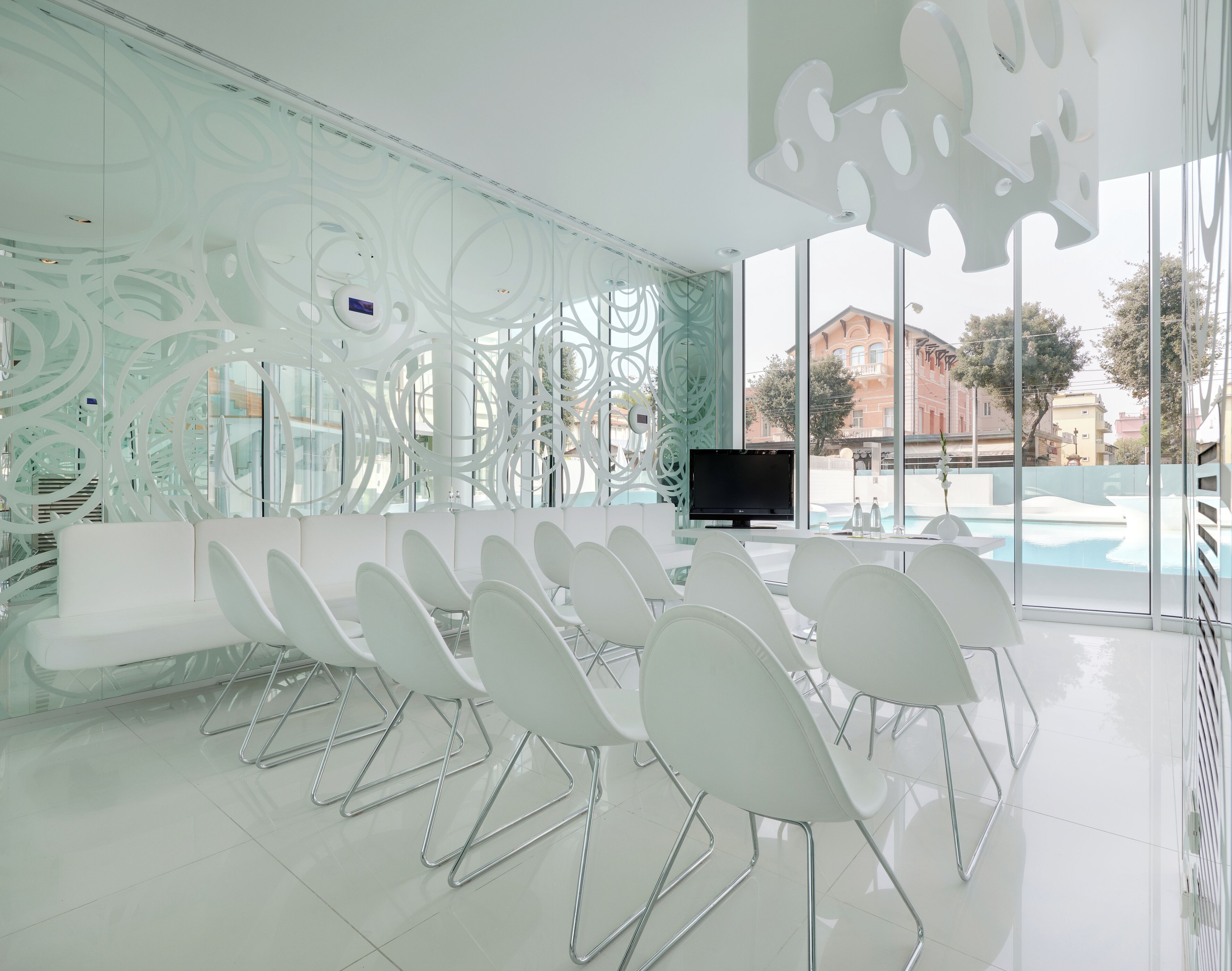 Une salle de conférence moderne, dotée de chaises blanches, d’un téléviseur à écran plat et d’un mur en verre offrant une vue sur une piscine et des bâtiments.