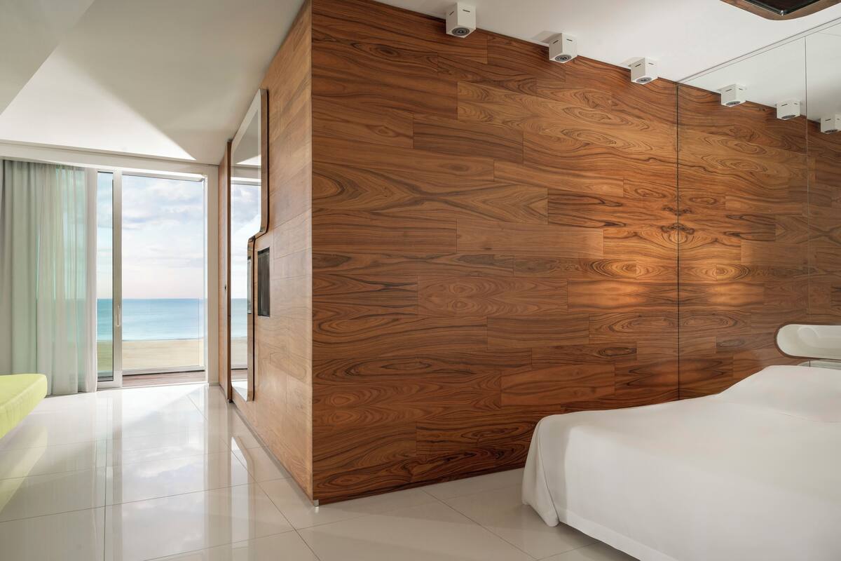 A modern bedroom with a large wooden panel wall, a bed, and a view of the beach through a glass door.