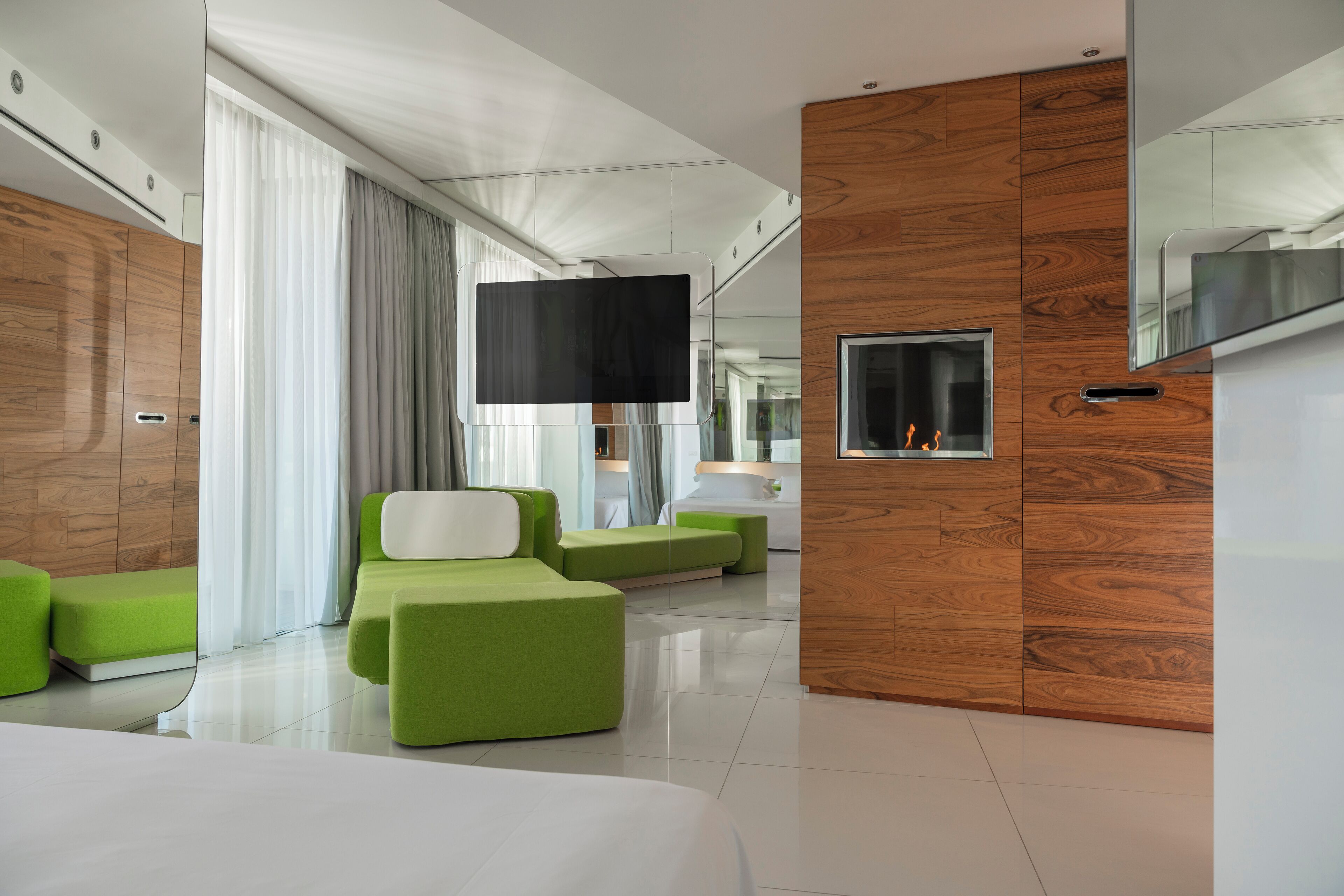 Una moderna camera d'hotel con un letto grande, un'area salotto verde, pannelli di legno, una televisione a schermo piatto e un caminetto.
