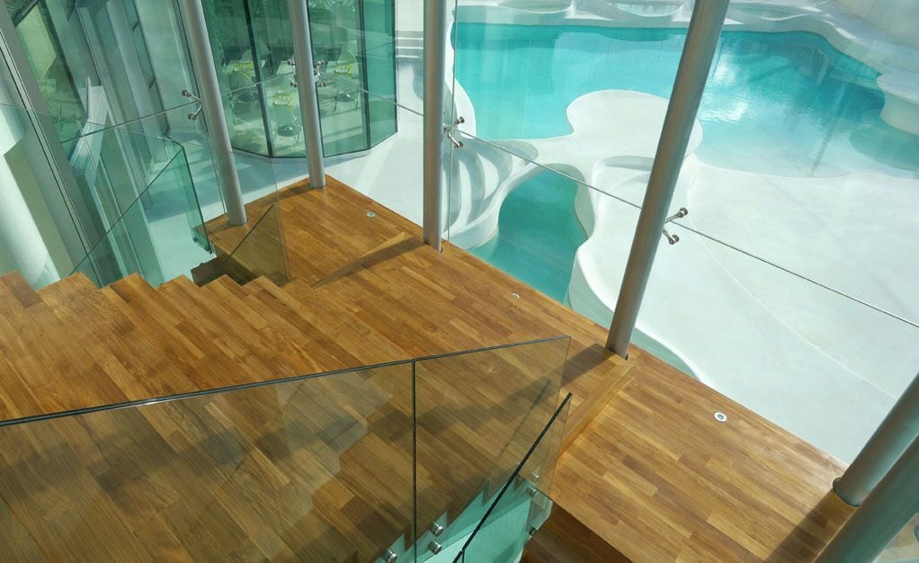Um interior moderno com escada de madeira, guarda-corpos de vidro e área de piscina visível através de grandes janelas.